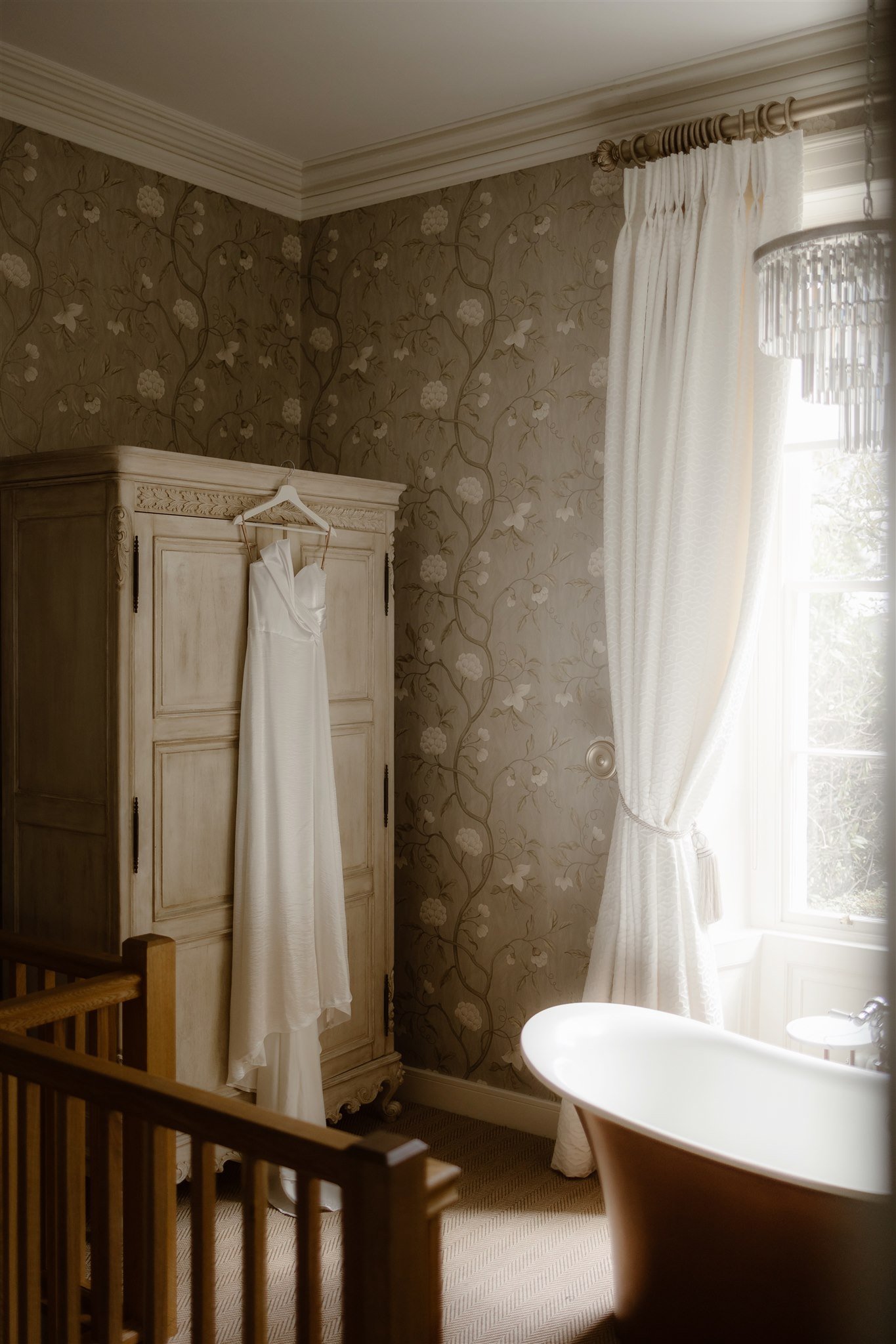 A white dress hangs on an antique wooden wardrobe in a softly lit, elegant bathroom at Town Head Estate, with floral wallpaper, white curtains, and a freestanding bath near a window.