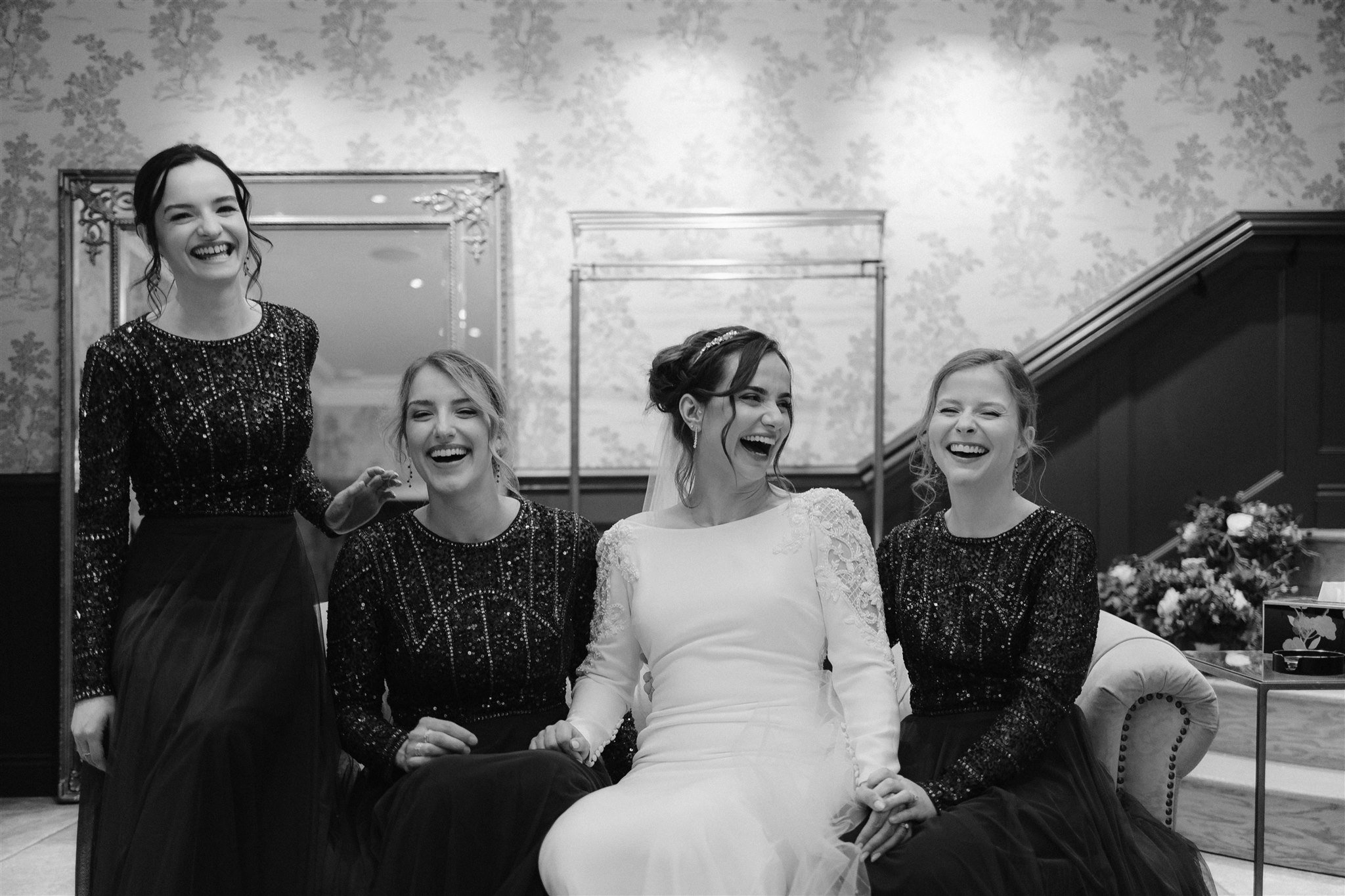 Four women sit and laugh together indoors at a Town Head Estate wedding. The woman in the centre wears a white dress, while the others wear matching dark, sparkly dresses. Ornate wallpaper, mirrors, and a staircase complete the elegant scene.