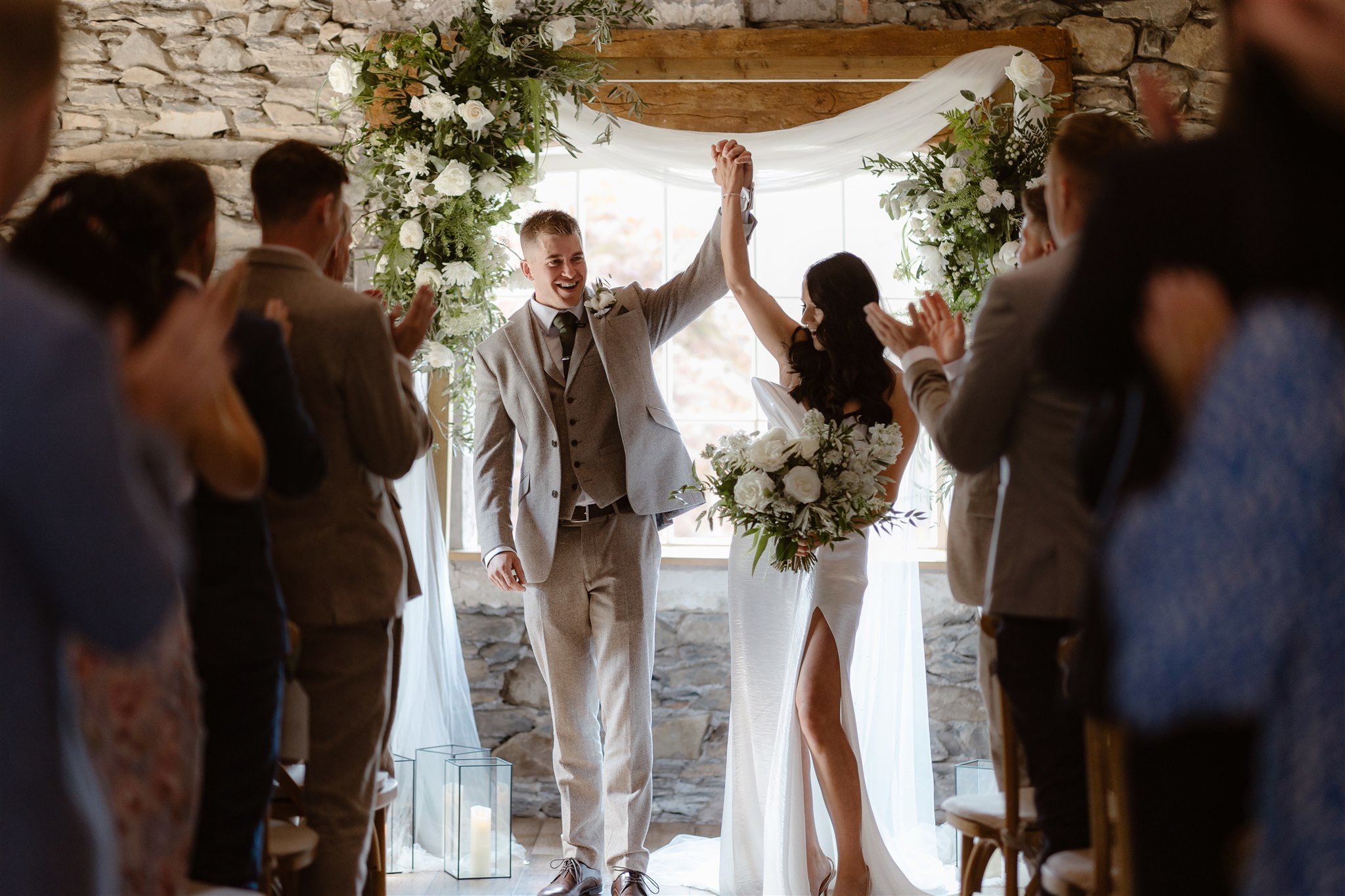 A newlywed couple stands at the altar during their Town Head Estate Wedding, holding hands triumphantly as guests applaud. Surrounded by greenery and flowers against a stone wall, they smile joyfully—the bride in white, the groom in a light suit.