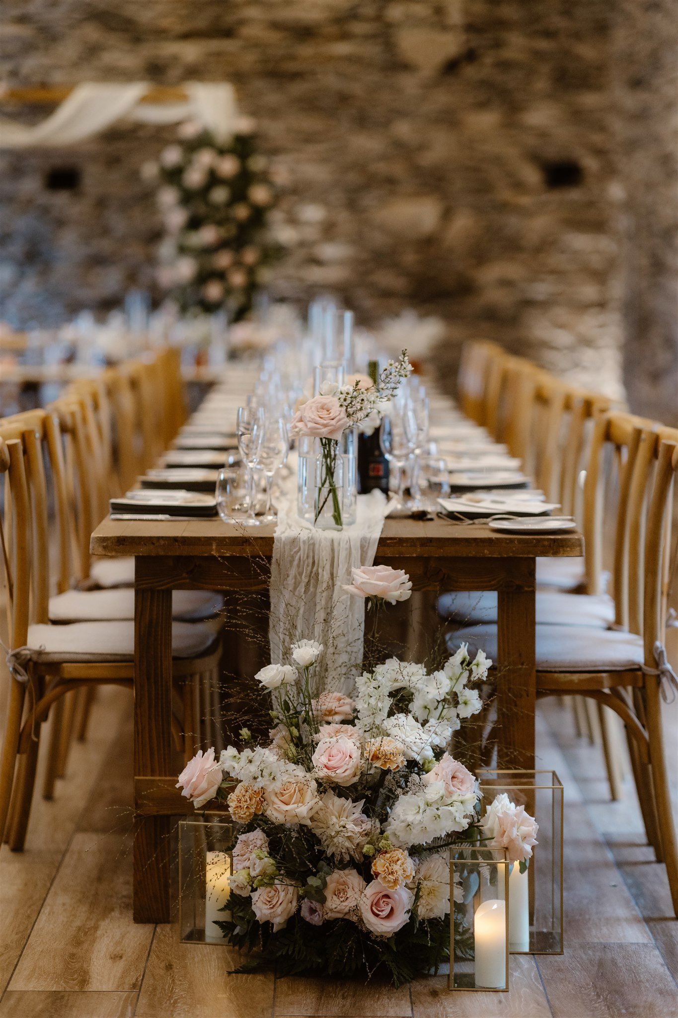 A long wooden table set for a formal Town Head Estate Wedding is decorated with glassware, plates, a lace runner, and pink and white floral arrangements. Wooden chairs line both sides, with candles and flowers adorning the table’s end.