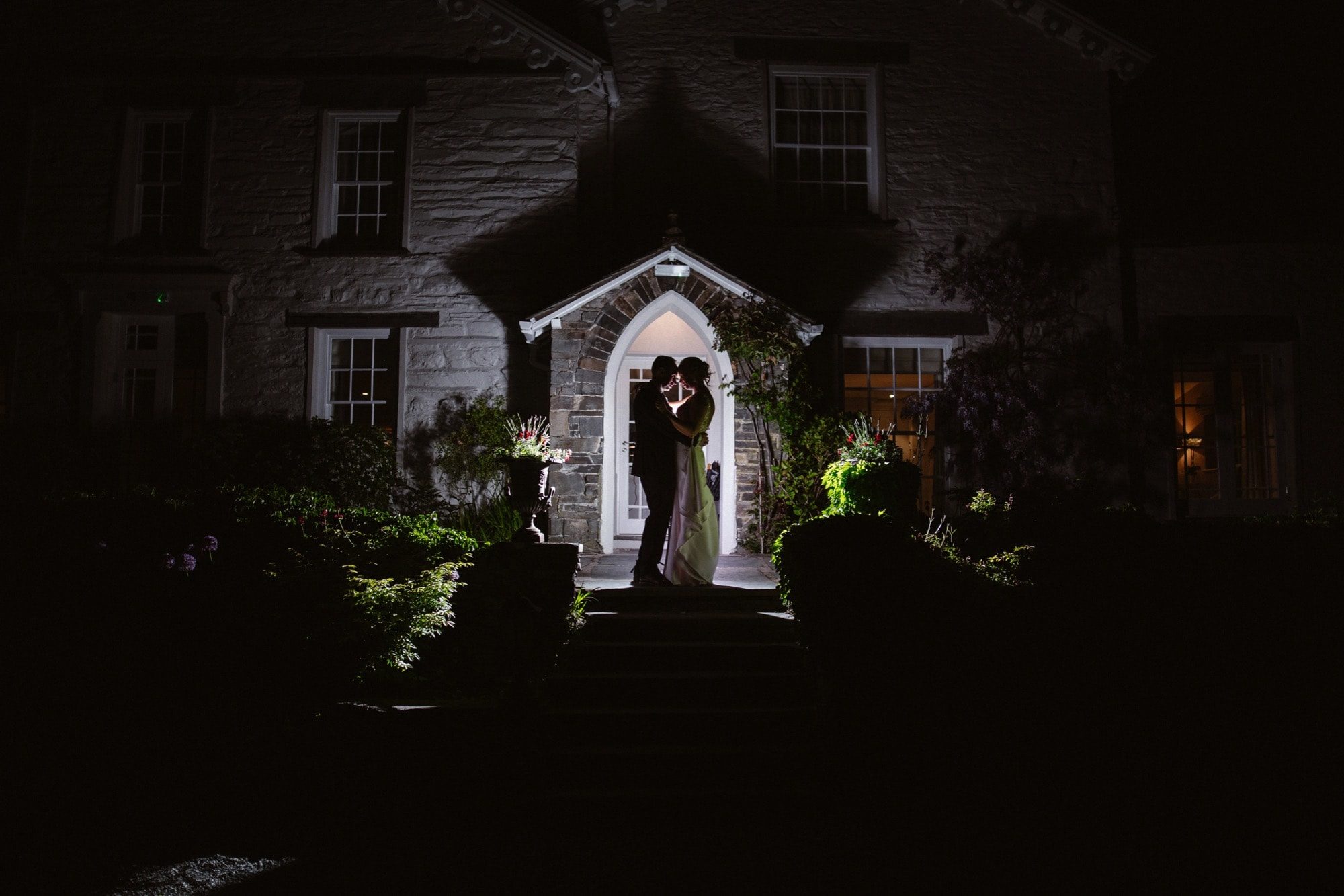 A couple stands embraced in the illuminated doorway of The Samling Hotel at night, framed by shadows and greenery, creating a romantic and dramatic silhouette.
