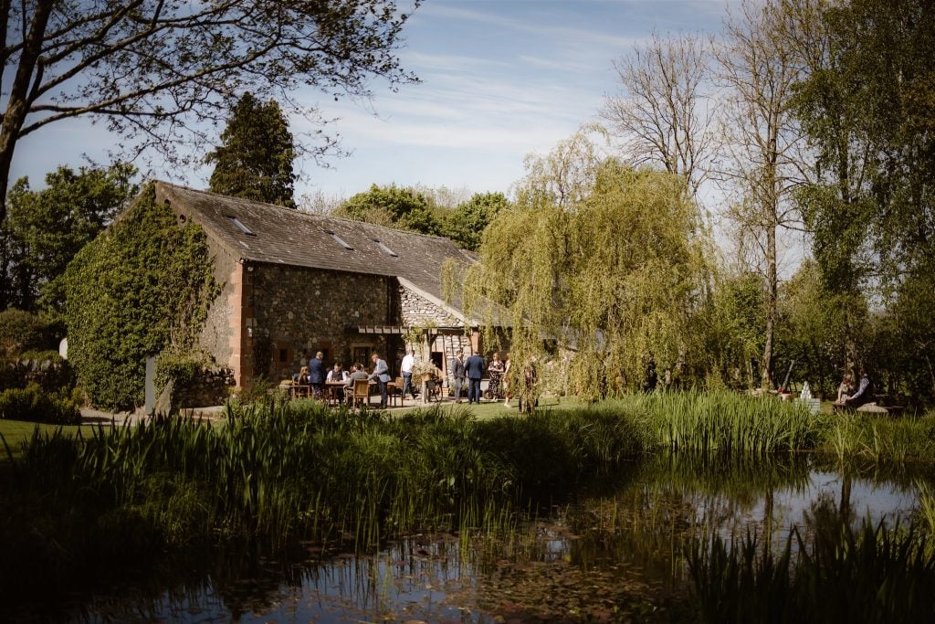 A rustic stone house, known as Low Hall, sits beside a pond surrounded by lush greenery and trees. Several people are gathered outside, enjoying the sunny weather and outdoor seating in this peaceful garden setting.