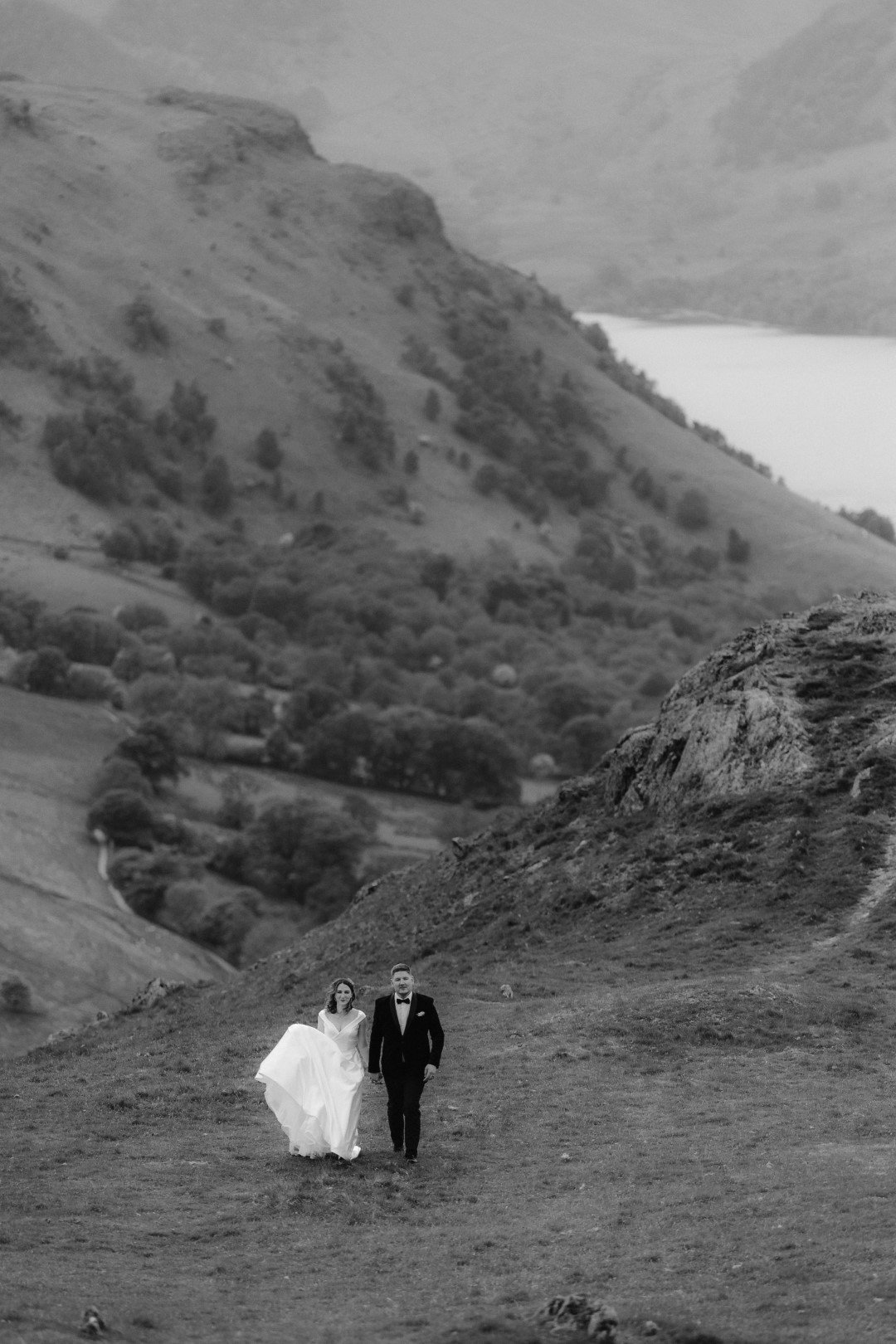 A bride and groom walk together up a grassy hillside near The Samling Hotel, surrounded by rolling hills and a distant lake, in a misty, black-and-white landscape.