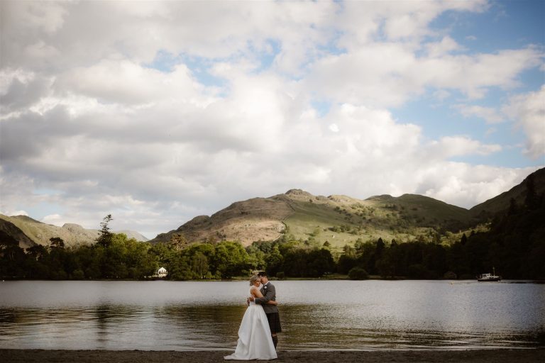 A newlywed couple embrace by a calm lake with lush green hills and partly cloudy skies, finding their own paradise in Another Place. The bride wears a white gown, and the groom wears a kilt.