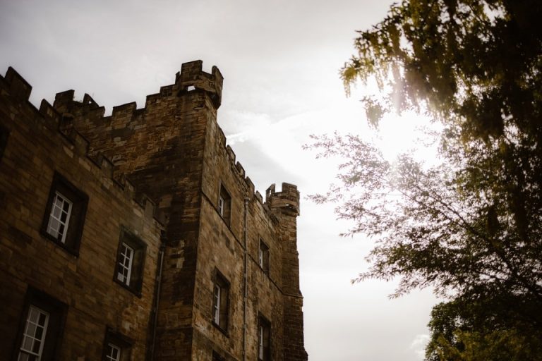 A stone castle with battlements, partially in shadow, rises against a bright sky as sunlight streams through tree branches—evoking the mysterious beauty of Another Place.