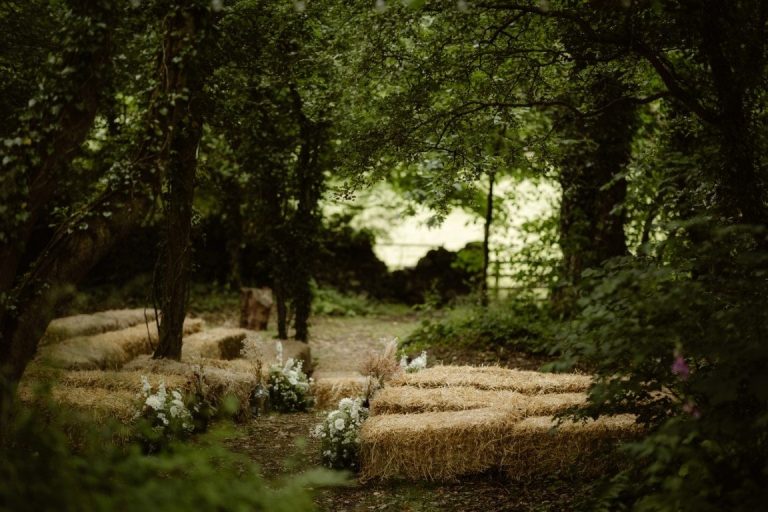 Outdoor woodland setting at Castletown Woodland Weddings, with rows of hay bales arranged as seating, decorated with white flowers, creating a natural aisle under lush green trees.