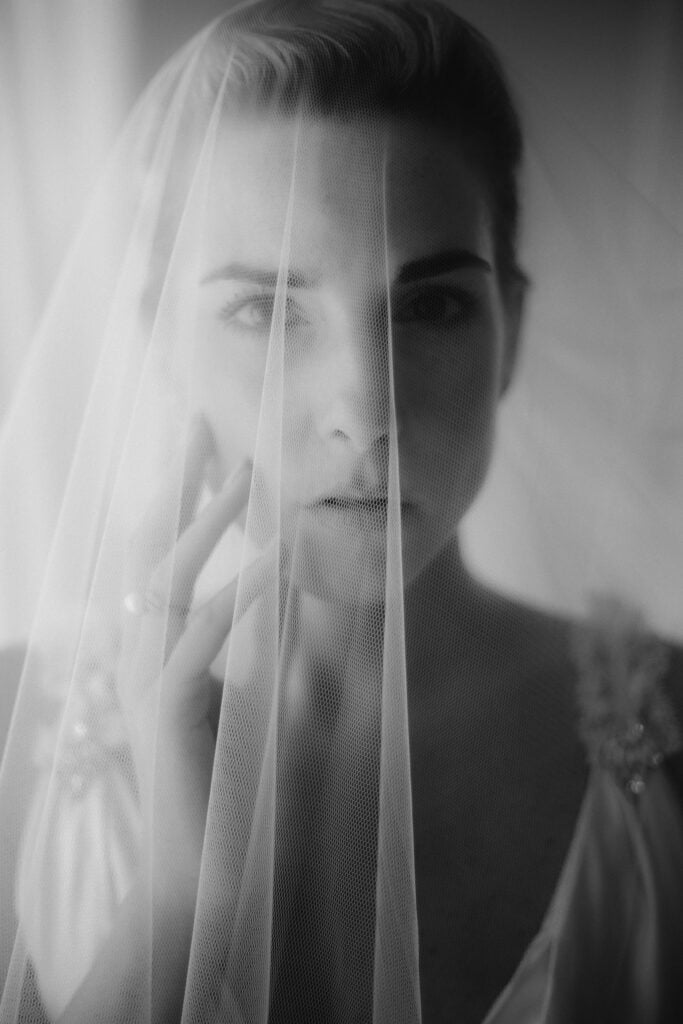 A woman in a wedding dress stands behind a delicate veil at The Samling Hotel. Shown in black and white, she gazes softly at the camera, gently touching the veil with her fingers.