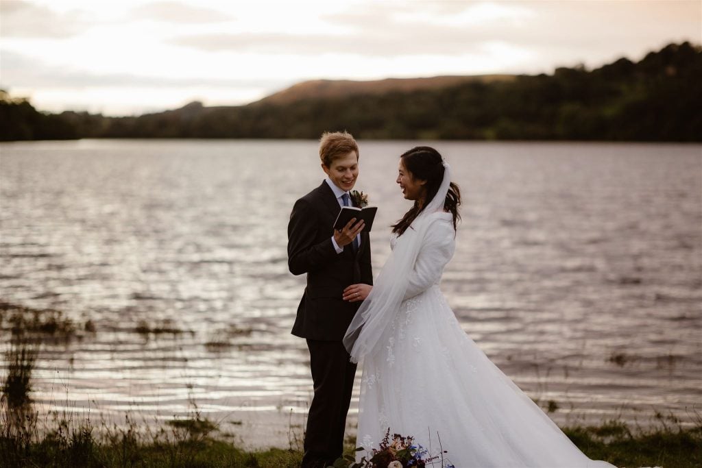 A couple stands by a lake at sunset near Low Hall; one in a suit reads from a book, while the other, in a white wedding dress, smiles and listens. Trees and hills appear in the background.
