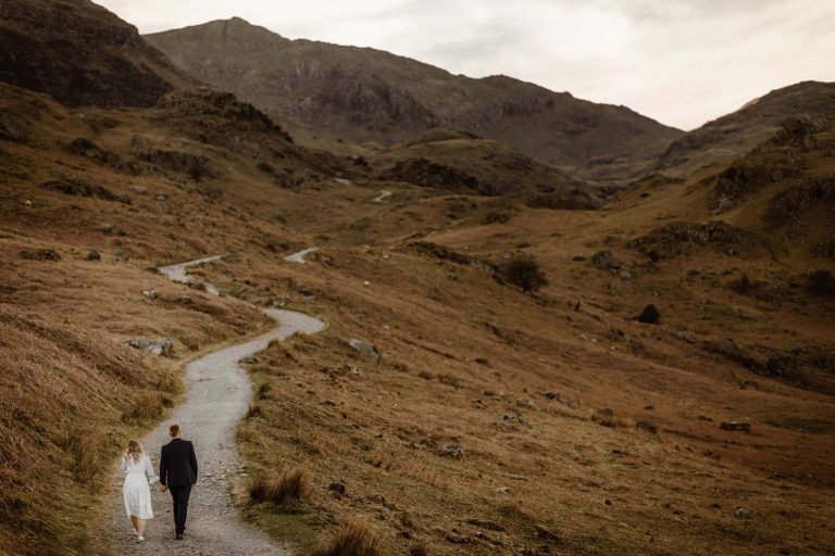 A couple walks hand-in-hand along a winding path through the vast, hilly landscape near The Copper Mines & Lakes Cottages, surrounded by brown grass and rocky terrain under an overcast sky.