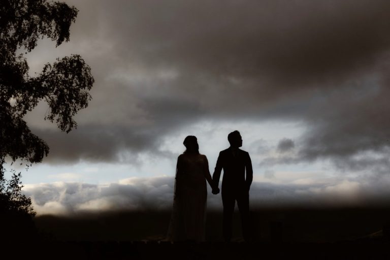 Silhouette of a couple holding hands, standing outdoors under a dramatic, cloudy sky with a tree on the left and distant hills in the background near Lodore Falls Hotel & Spa.