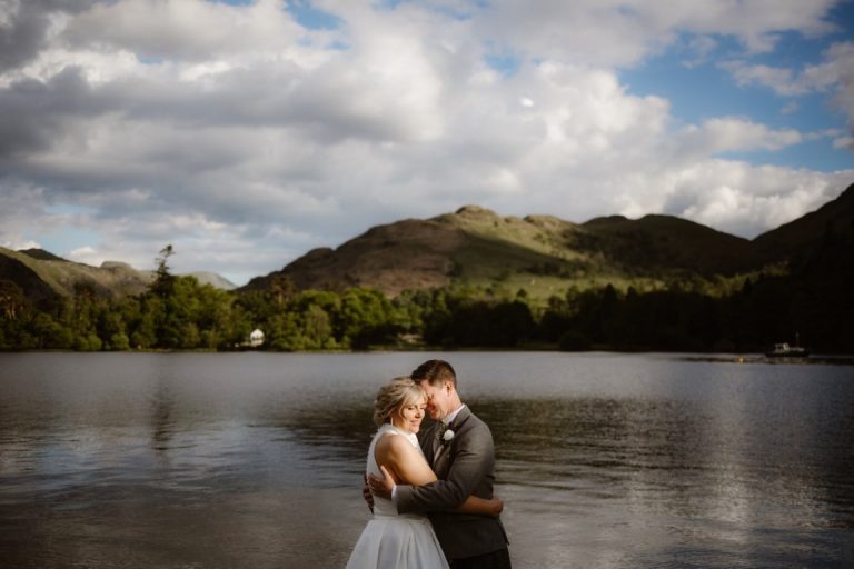 A bride and groom in wedding attire embrace by a calm lake near MacDonald Leeming House, with green hills and a cloudy blue sky in the background, creating a romantic and serene outdoor scene.
