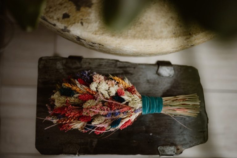 A bouquet of dried, colourful flowers from Storrs Hall, wrapped with a teal ribbon, rests on a rustic wooden surface. The flowers display vibrant shades of red, white, purple, and pink when viewed from above.