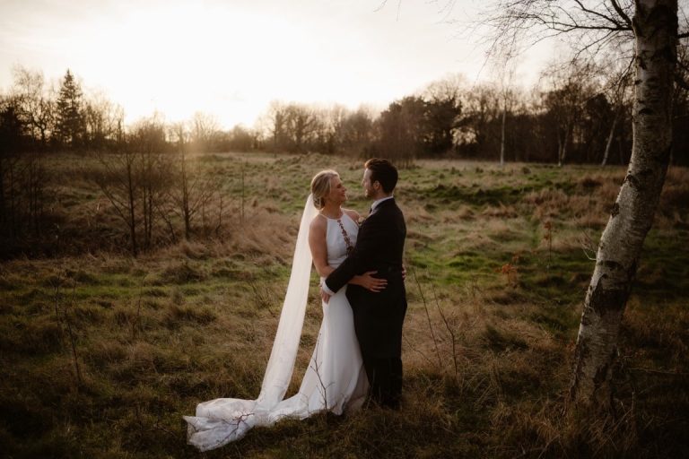 A bride and groom stand together in a grassy field at sunset at The Garden at Eden, gazing at each other lovingly. The bride’s long veil and white dress glow in the soft sunlight, whilst trees frame their romantic moment.