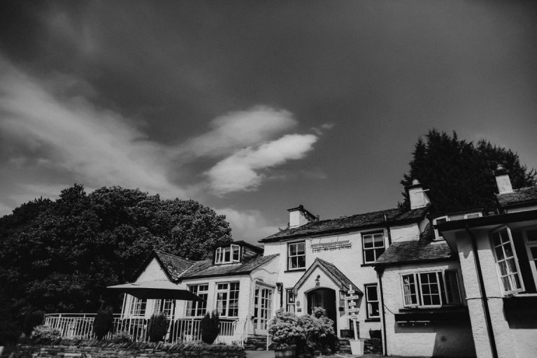 Black and white photo of The Wild Boar, a large house with white walls and multiple chimneys, surrounded by trees. The sky is partly cloudy, and the building features various windows and an outdoor seating area.