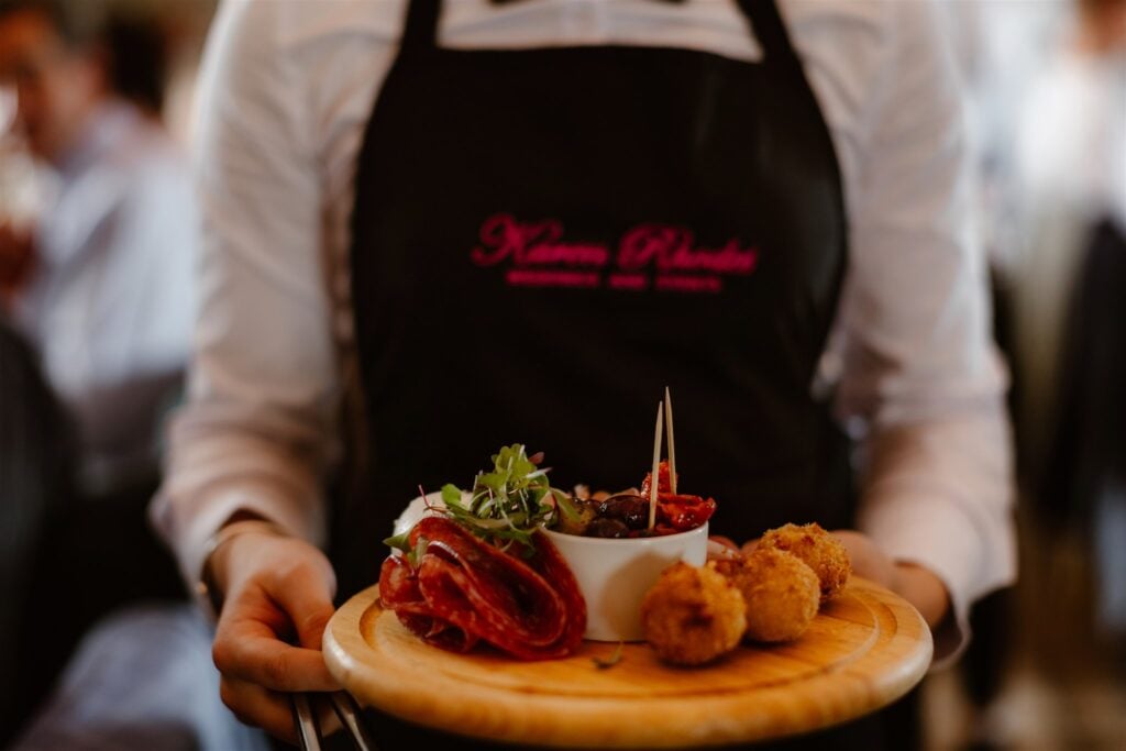 A waiter in a black apron presents a wooden platter with cured meats, dip, olives, tomatoes, greens, and breaded bites on skewers at a Town Head Estate Wedding. The background is softly blurred.