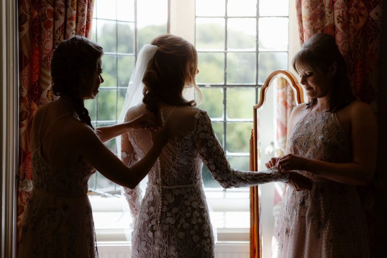 A bride in a lace wedding dress stands by a bright window, as two women in light dresses help her get ready, adjusting her dress and veil. Warm light streams in, illuminating the scene.