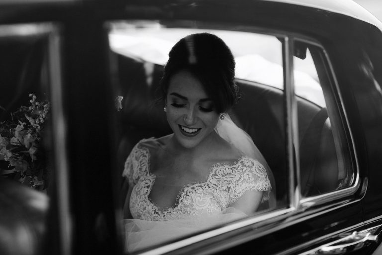 A bride in a lace wedding dress sits smiling inside a car, viewed through the window. The black and white image gives the scene an ethereal feel, as if she’s dreaming of Another Place beyond her serene expression and elegant hairstyle.