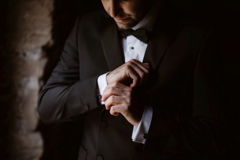 A man dressed in a black dinner jacket adjusts his cufflinks in dim lighting, as if preparing for Another Place, with only his torso and lower face visible, creating an elegant and refined atmosphere.