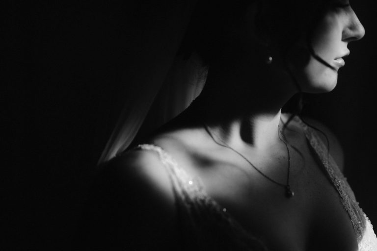 A black and white close-up photo of a bride in soft lighting at Three Hills Barn, showing her shoulder, neckline, and part of her face in profile. She wears a necklace, veil, and lace wedding dress. Shadows create a dramatic, elegant mood.