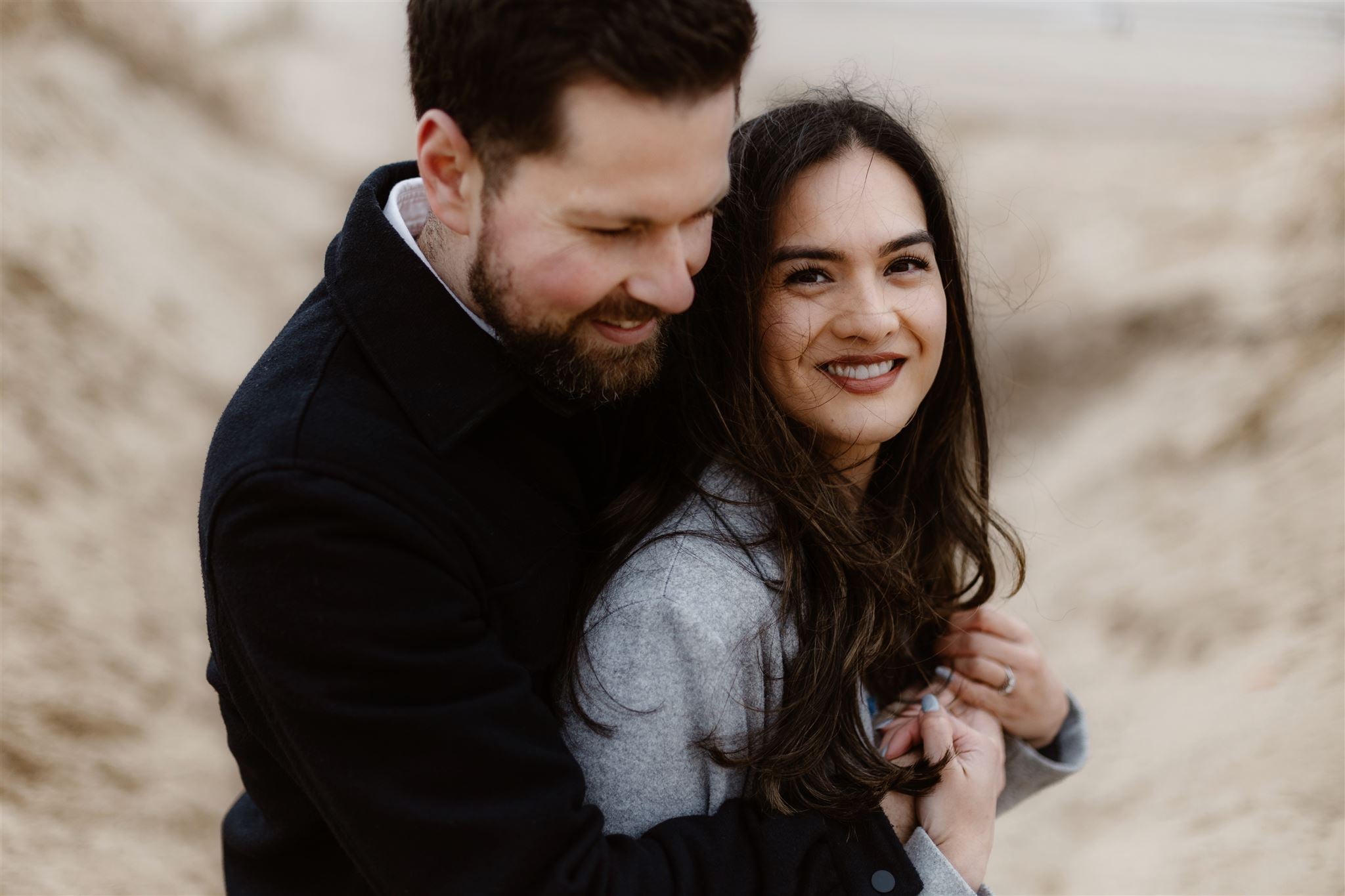 A man embraces a smiling woman from behind as they stand outdoors on a sandy beach. Both are wearing coats, and the woman looks at the camera while the man looks down, smiling.