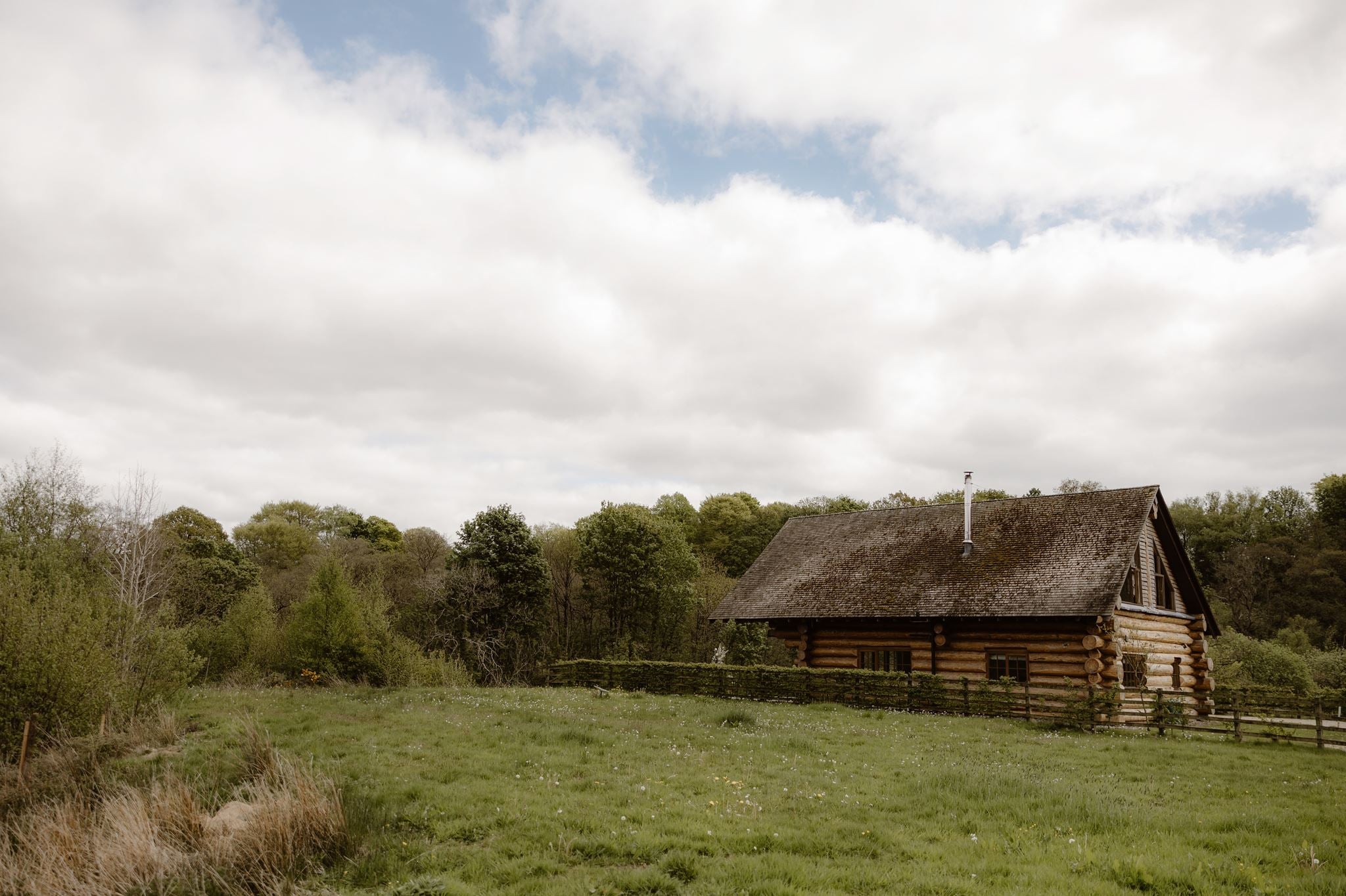 A rustic log cabin with a wooden roof sits in a grassy field in front of a line of trees under a cloudy sky.
