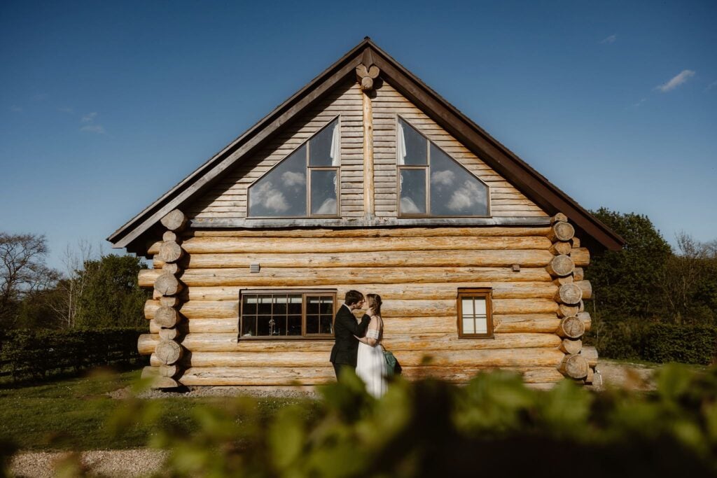 A couple stands kissing in front of a large log cottage at Hidden River Cabins.